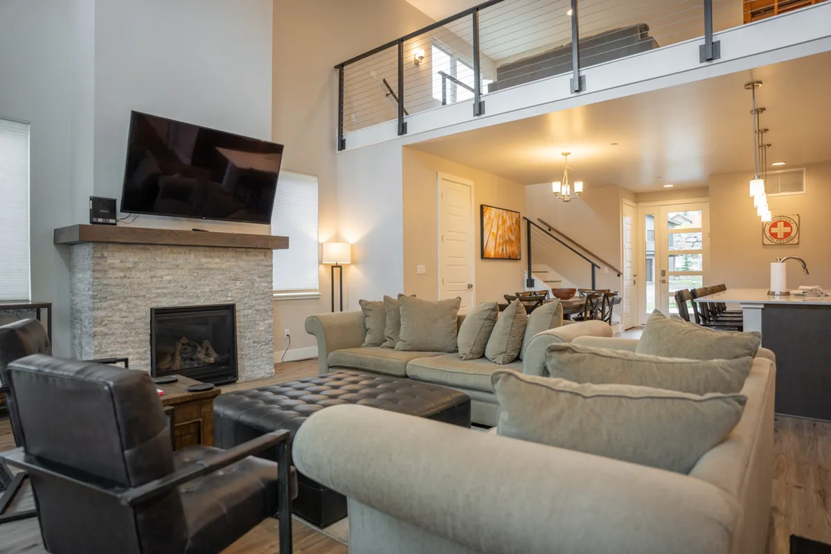 The image showcases a spacious, warmly lit living and dining area featuring a plush sofa, a crackling fireplace, a large television, and an inviting dining table—perfect for gatherings. The mood is cozy and welcoming, ideal for families and groups to relax, share meals, and enjoy each other's company after a day on the slopes or hiking nearby trails.
