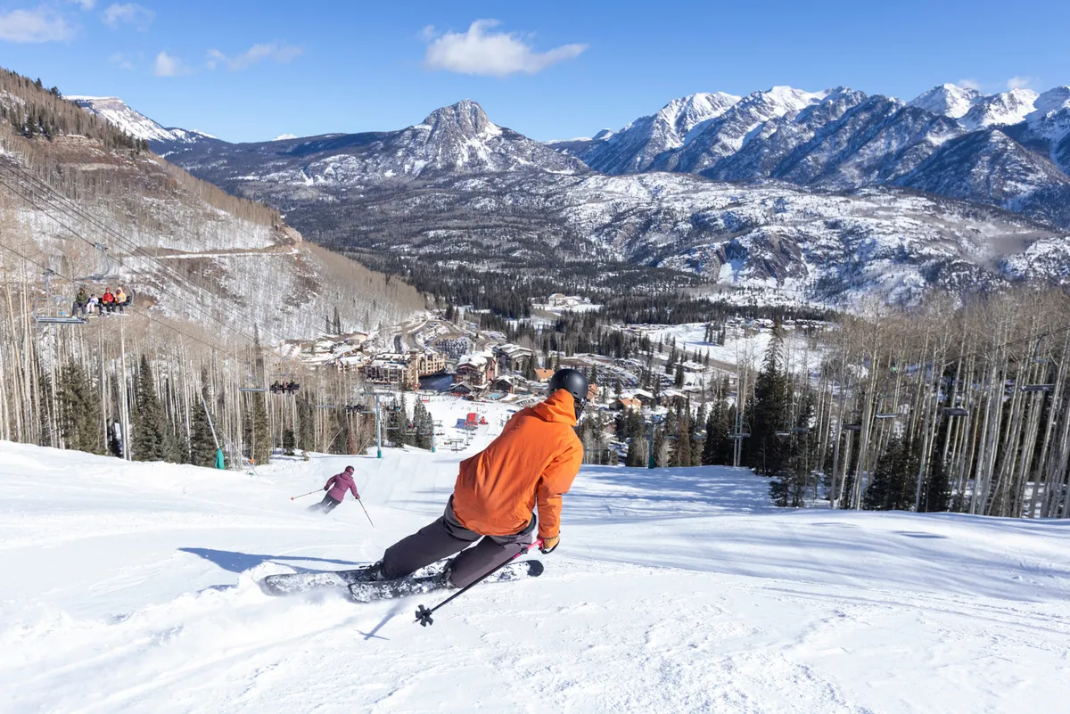 A skier dressed in a bright orange jacket descends a snow-covered slope with a ski lift and dramatic mountain range in the background. The wintry scene is energetic and exhilarating, embodying the excitement of ski season at the resort.