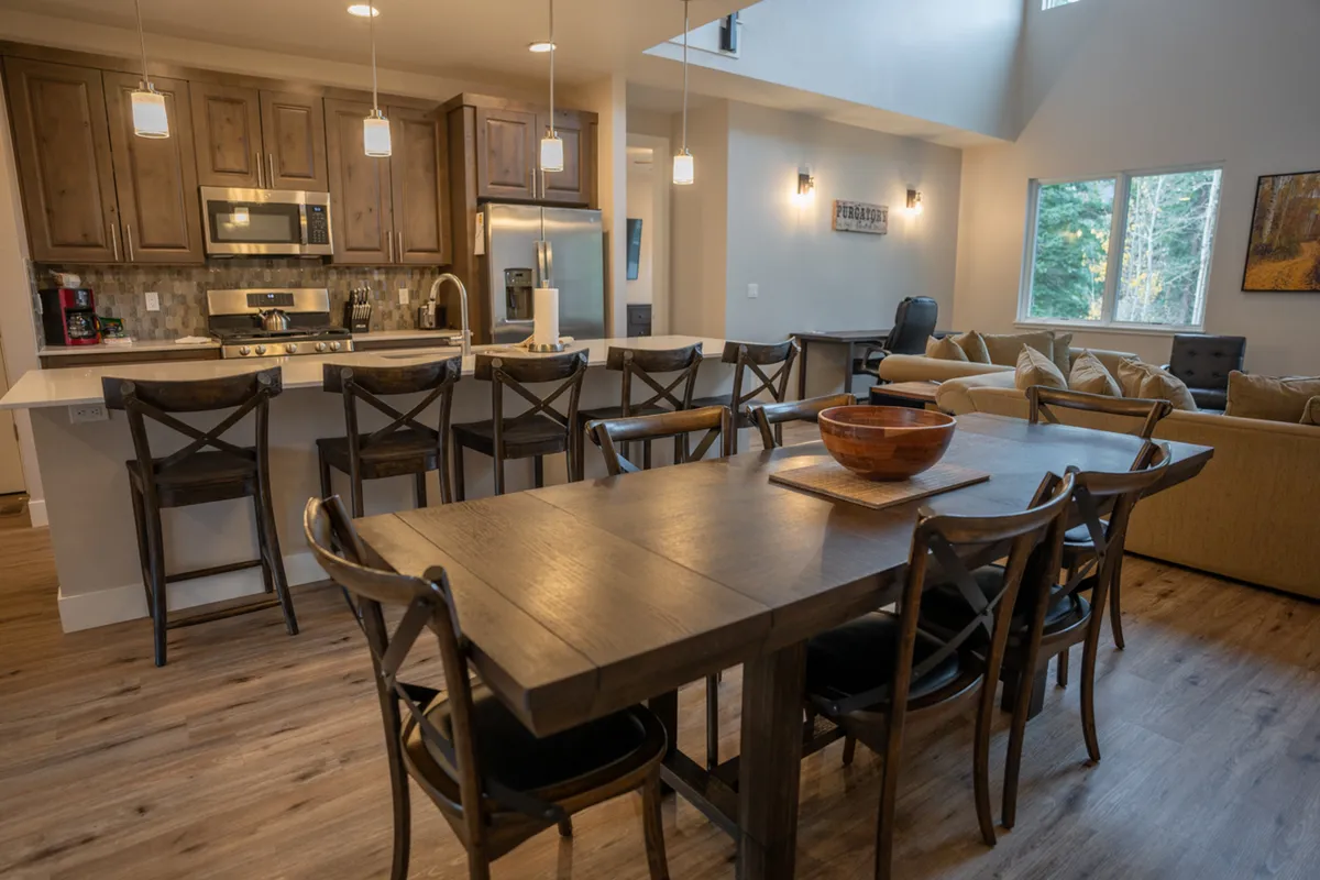 The image highlights an elegant dining table, kitchen island, and comfortable sofa area, all illuminated by natural light streaming through large windows. The scene evokes shared meals, celebrations, and relaxation with loved ones.