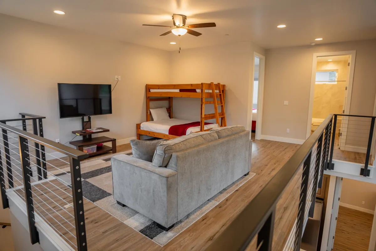 A cozy loft area featuring a queen/twin bunk bed and a sofa, with a television for entertainment. The space is arranged to host both children and extra guests, offering a playful yet practical sleeping and lounging solution.