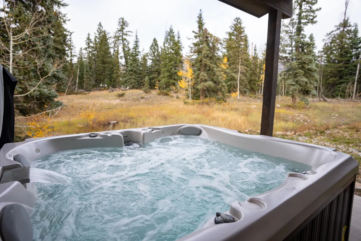 A peaceful exterior view of a hot tub set amidst natural surroundings, with trees and an open field visible. The area is tranquil and ideal for relaxation, and the scene hints at both sunset and stargazing possibilities. The hot tub sits close to nature, creating a direct connection to the outdoors.