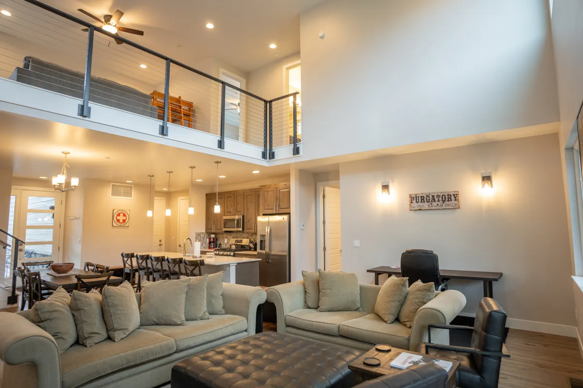The photo showcases a spacious, open-concept great room with a large sectional sofa, a visible open kitchen, and a sizable dining area featuring a wooden table. The staircase to the loft and ample natural light create a comfortable, communal atmosphere.
