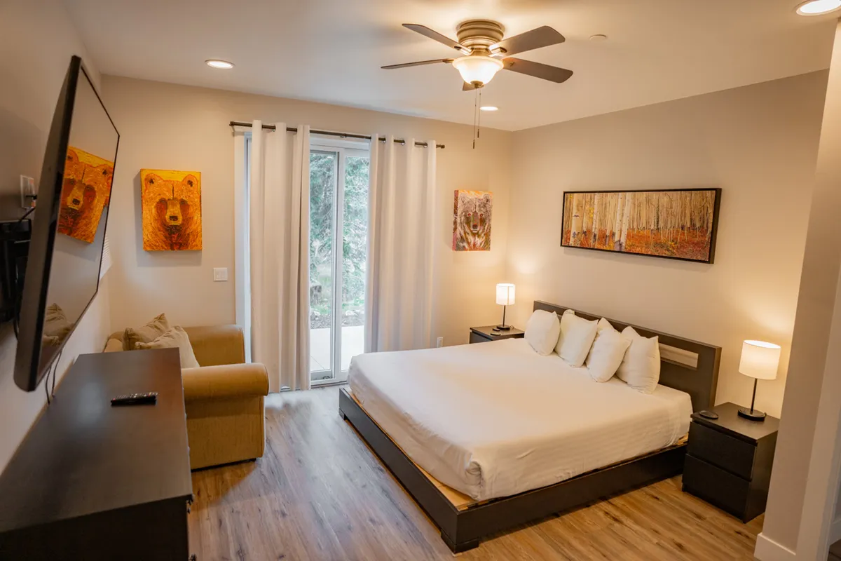 A spacious bedroom with a large king bed dressed in crisp white linens, accented by nature-inspired artwork depicting bears and autumn trees. A comfortable sofa seating area and a wall-mounted television provide relaxation options, while large windows allow natural light to fill the space.
