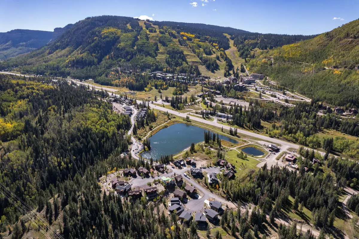 A sweeping aerial view encompassing mountain slopes, a lakeshore, forested areas, and a residential neighborhood. The landscape is alive with adventure opportunities, hinting at hiking trails, skiing runs, and tranquil lakeside settings.