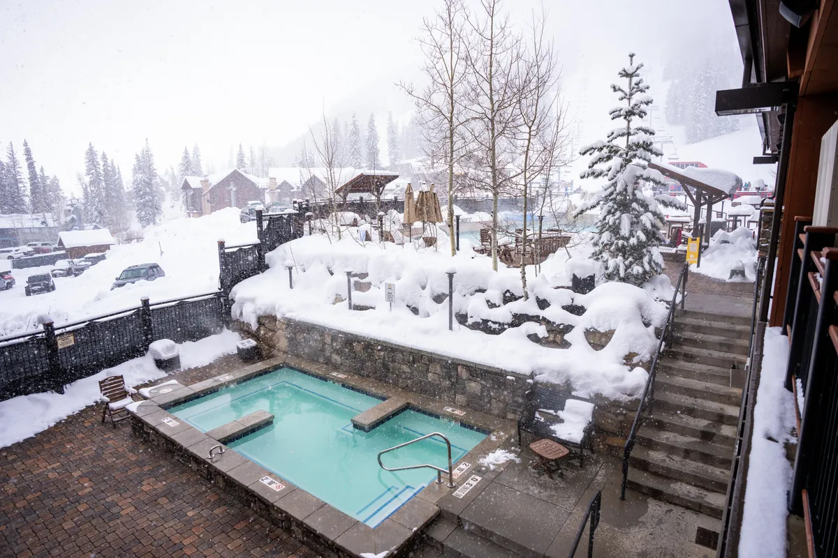 An exterior shot featuring a private hot tub surrounded by snow-covered landscapes and trees. The hot tub is situated on a deck area, with stairs leading up and a cozy, tranquil ambiance. People are shown relaxing in the hot tub, enjoying the picturesque winter scenery.