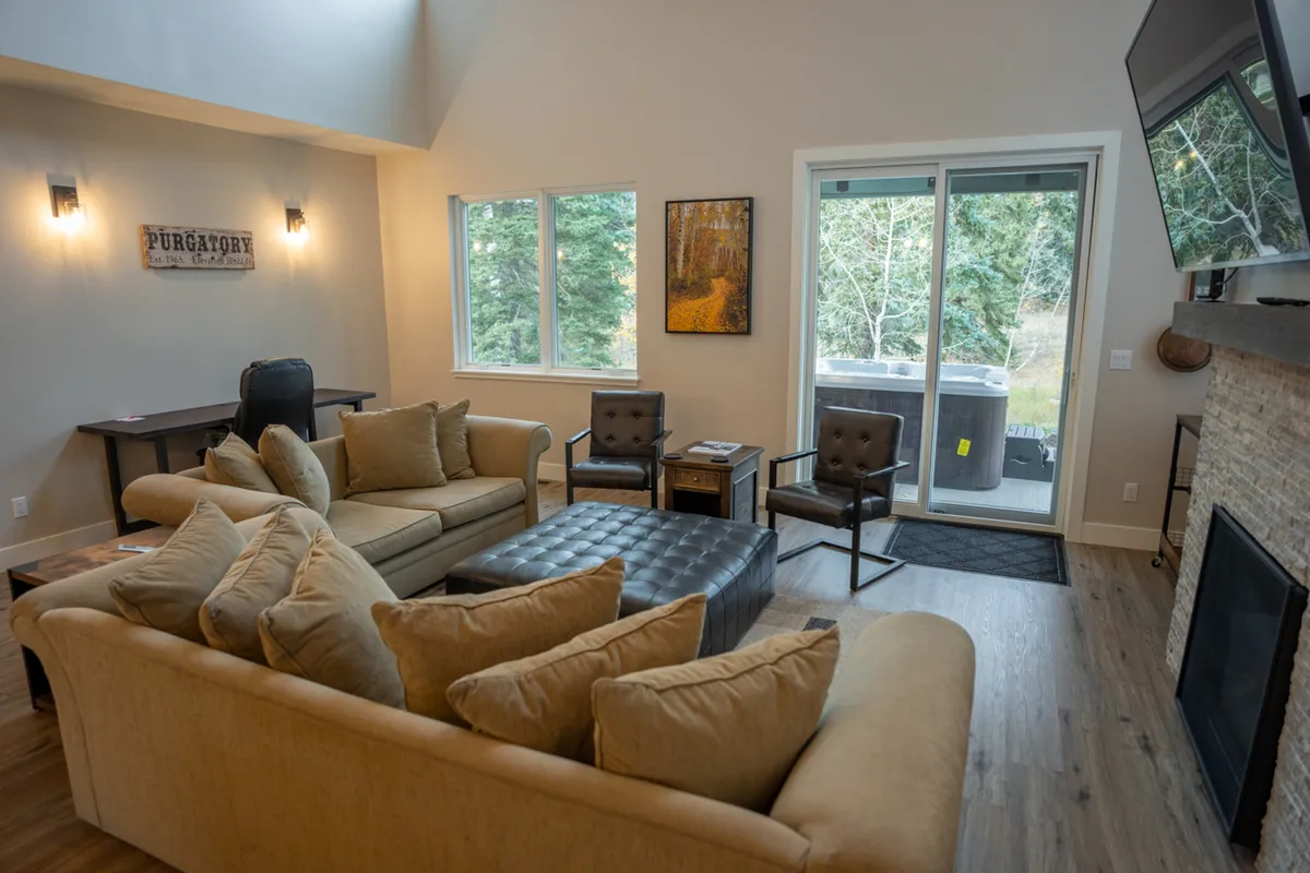 A warm and inviting living space centers on a comfortable sofa facing a gas fireplace, with large windows letting in mountain light and an art piece adorning the wall. The coffee table suggests a space for relaxed evenings or board games.