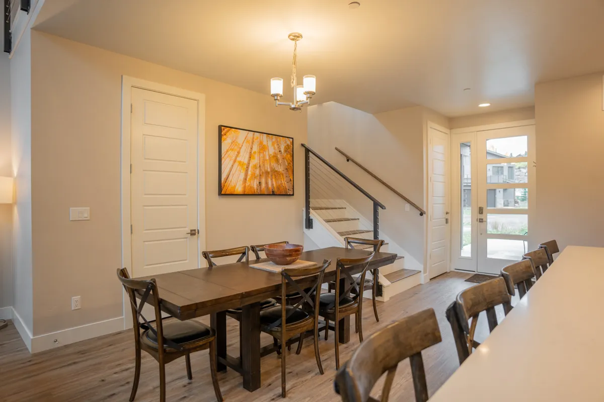 An inviting dining area with a large table set beneath a stylish chandelier, complemented by expansive windows and warm wood accents. The space is designed for memorable group meals, socializing, and enjoying morning coffee with a mountain view.