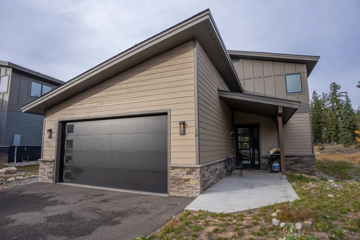 A welcoming exterior view of the home, featuring the garage door, an entrance porch, and a sturdy stone foundation. The porch area is perfect for gatherings or enjoying nature, and the surrounding greenery sets a tone of tranquility and accessibility.