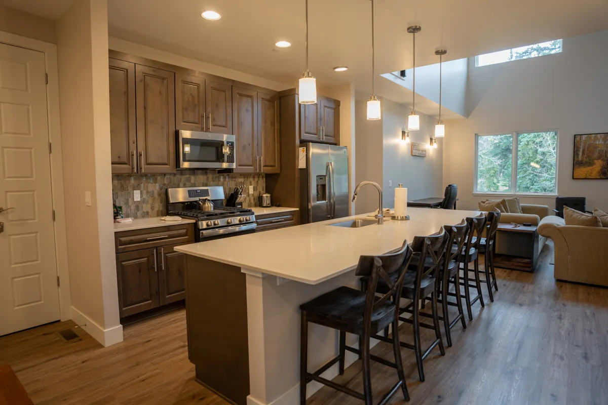 A bright kitchen island with stainless steel appliances is the focal point, bathed in natural light from large windows. There's a cozy adjacent seating area, inviting for conversation or relaxation while cooking.