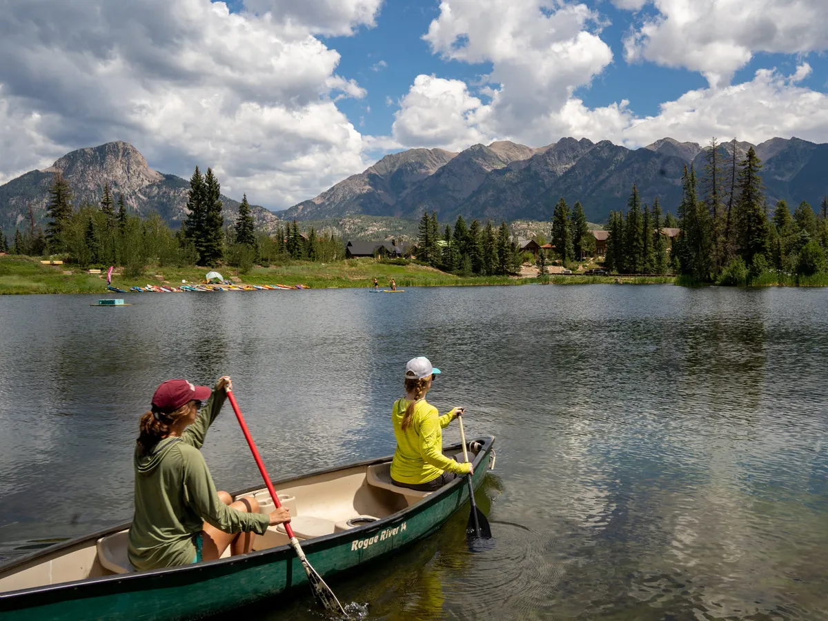 Two women paddle a canoe on a glassy lake with mountains rising in the background, surrounded by trees and nearby houses. The calm, reflective water suggests peaceful relaxation while highlighting the recreational possibilities of Twilight Lake.