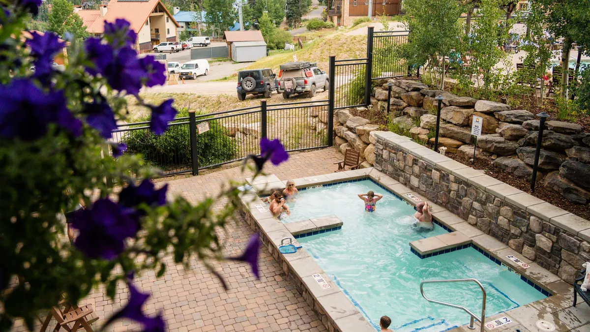 An inviting outdoor hot tub surrounded by blooming flowers and a stone wall, with people relaxing and enjoying the sunset. The environment exudes a sense of relaxation and community after a day of adventure.